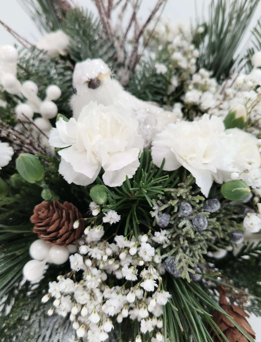 Snowy Perch floral arrangement with white carnations, baby’s breath, evergreen, pinecones, and a winter bird.
